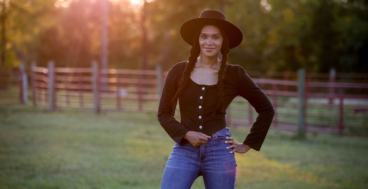 A photo of a woman wearing a black shirt, black felt hat, and blue jeans, posing in a green pasture.